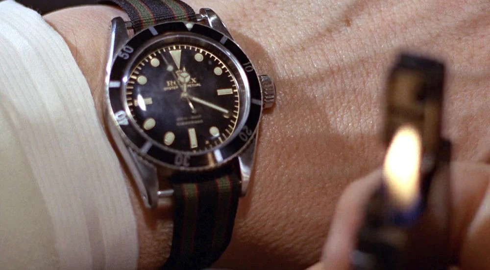 A Rolex Submariner mechanical watch is being worn on a persons wrist. A lighter is shown out of focus in the frame.
